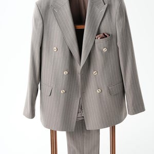 Puede incluir: Un traje gris con finas rayas, que incluye una americana cruzada y pantalones a juego, cuelga de un perchero de madera. La americana tiene botones dorados y un pa&ntilde;uelo de bolsillo. El traje se exhibe sobre un fondo blanco.