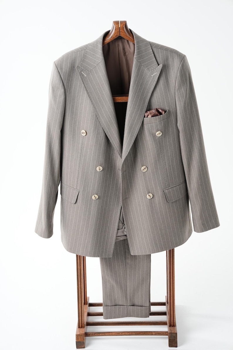 Puede incluir: Un traje gris con finas rayas, que incluye una americana cruzada y pantalones a juego, cuelga de un perchero de madera. La americana tiene botones dorados y un pa&ntilde;uelo de bolsillo. El traje se exhibe sobre un fondo blanco.