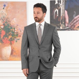 May include: A man in a gray suit, white shirt, and patterned tie stands in front of two paintings. The suit is a solid gray color, and the tie is a lighter shade. One painting depicts a vase of flowers, and the other shows hands sewing.