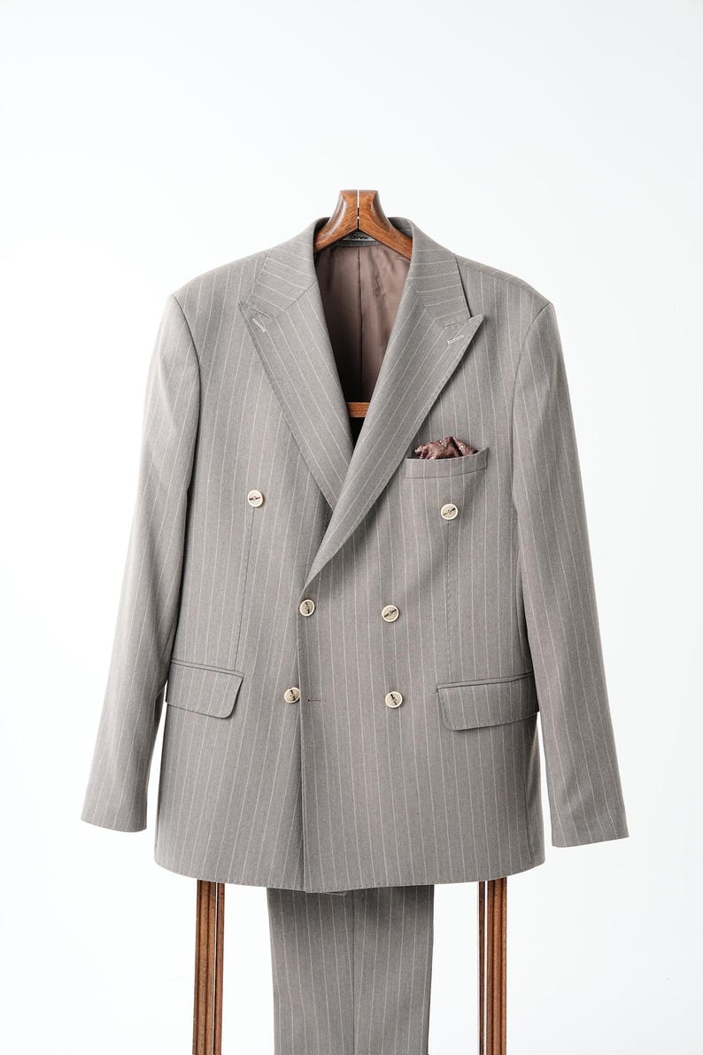 Puede incluir: Un traje gris de doble botonadura con finas rayas blancas, botones dorados y un pa&ntilde;uelo de bolsillo. El traje se exhibe en una percha de madera, mostrando un dise&ntilde;o cl&aacute;sico y a medida. El conjunto incluye una chaqueta y un pantal&oacute;n a juego.