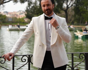 May include: A cream-colored double-breasted blazer with a black bow tie and black pants. The blazer has a satin lapel and is worn over a white dress shirt. A silver watch is visible on the wrist.