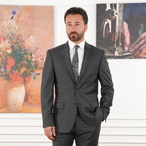May include: A charcoal gray suit with a white dress shirt and patterned tie. The suit jacket has two buttons and a pocket on each side. The background includes two paintings, one of a floral arrangement and the other of a person sewing.