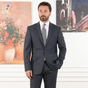 May include: A man in a dark gray suit, white shirt, and patterned tie stands in front of two paintings. The suit has a two-button closure and a tailored fit. The background includes a floral still life and a sewing machine.