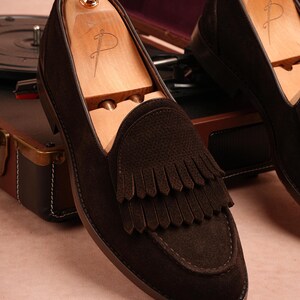 May include: Dark brown suede loafers with a fringed design. The shoes are shown with wooden shoe trees. The loafers have a leather sole and stitched detailing. A vintage record player and leather case are in the background.