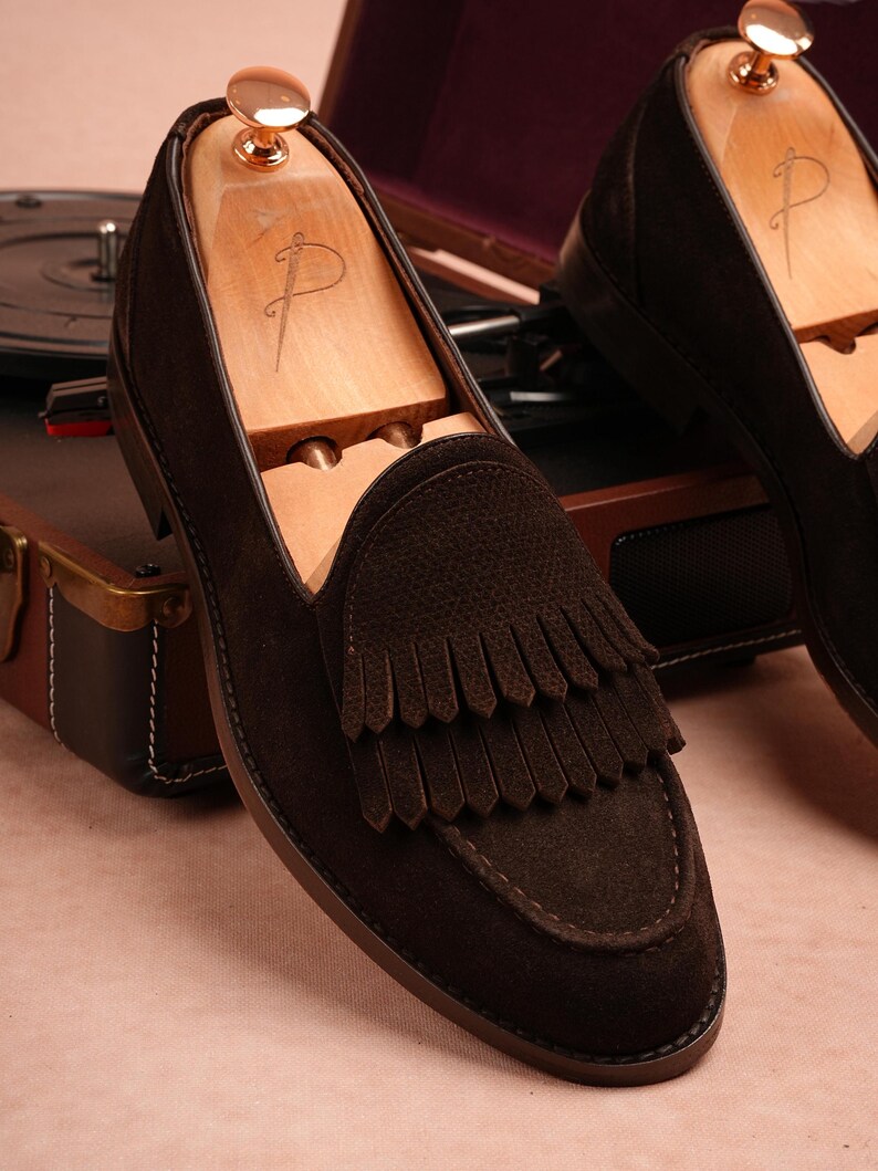 May include: Dark brown suede loafers with a fringed design. The shoes are shown with wooden shoe trees. The loafers have a leather sole and stitched detailing. A vintage record player and leather case are in the background.