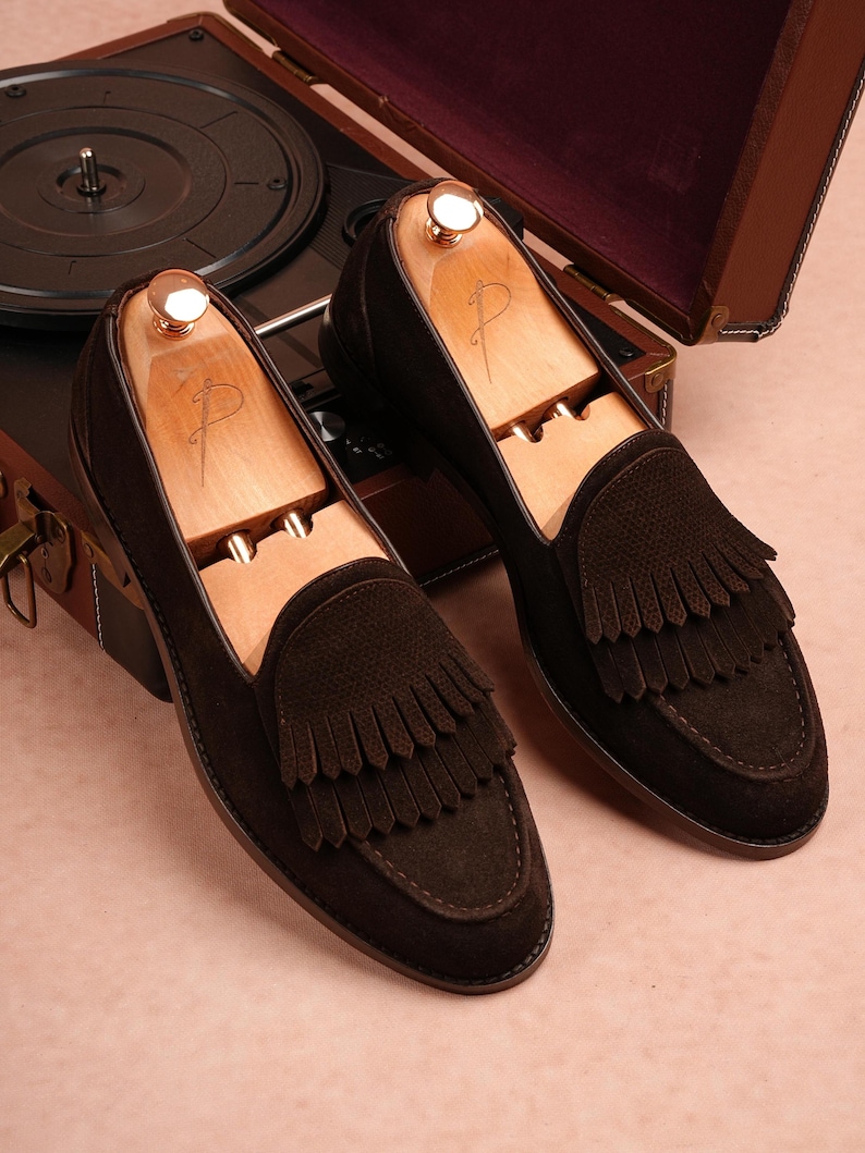 May include: Dark brown suede loafers with fringe detailing, presented with wooden shoe trees. The shoes are set against a vintage record player and a brown leather case.