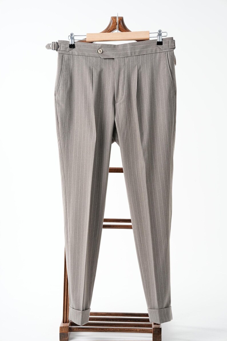 Puede incluir: Pantalones grises de raya diplom&aacute;tica colgados de una percha de madera. Los pantalones tienen cierre de bot&oacute;n, pliegues y dobladillos con vuelta. Los pantalones se exhiben en un perchero de madera sobre un fondo blanco.