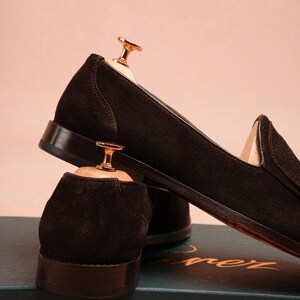May include: A pair of dark brown suede loafers with leather soles, displayed with wooden shoe trees. The shoes are set against a soft pink backdrop. The loafers have a classic design, suitable for formal or smart casual wear.