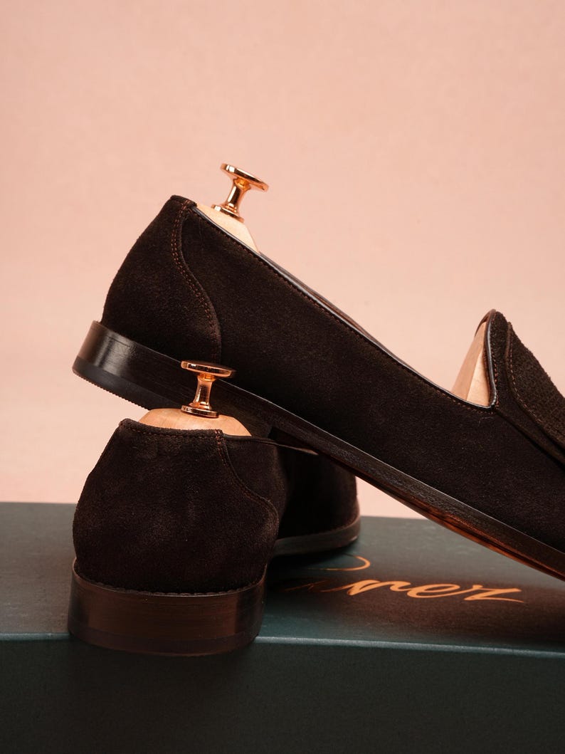 May include: A pair of dark brown suede loafers with leather soles, displayed with wooden shoe trees. The shoes are set against a soft pink backdrop. The loafers have a classic design, suitable for formal or smart casual wear.