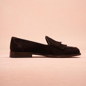 May include: A single dark brown suede loafer with a tassel detail. The shoe has a low heel and a classic, slip-on design. The shoe is set against a light pink background.