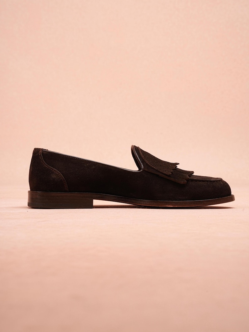 May include: A single dark brown suede loafer with a tassel detail. The shoe has a low heel and a classic, slip-on design. The shoe is set against a light pink background.