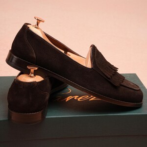 May include: Dark brown suede loafers with tassel detailing. The shoes are displayed on a teal box. Wooden shoe trees are inserted. The shoes are a classic style, suitable for various occasions.