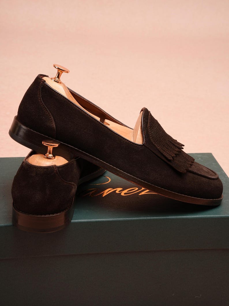 May include: Dark brown suede loafers with tassel detailing. The shoes are displayed on a teal box. Wooden shoe trees are inserted. The shoes are a classic style, suitable for various occasions.