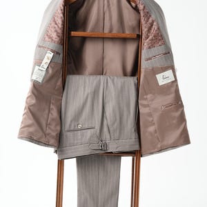 Puede incluir: Un traje gris a rayas finas, que incluye una chaqueta y un pantal&oacute;n, exhibido en una percha de madera. El interior de la chaqueta es marr&oacute;n claro con un forro estampado. Los pantalones tienen un dise&ntilde;o a rayas finas a juego y una hebilla. El traje est&aacute; sobre un fondo blanco.