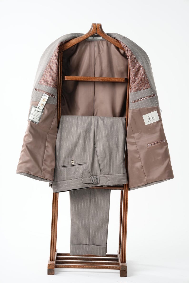 Puede incluir: Un traje gris a rayas finas, que incluye una chaqueta y un pantal&oacute;n, exhibido en una percha de madera. El interior de la chaqueta es marr&oacute;n claro con un forro estampado. Los pantalones tienen un dise&ntilde;o a rayas finas a juego y una hebilla. El traje est&aacute; sobre un fondo blanco.