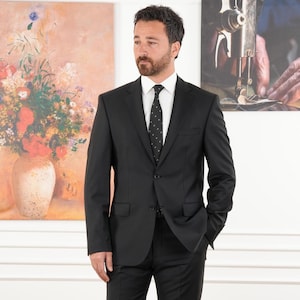 May include: A black suit with a white dress shirt and a black tie with white polka dots. The suit has a two-button closure and a notched lapel. The background includes two paintings, one with a floral still life and the other with a sewing machine.