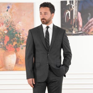 May include: A charcoal gray suit with a white dress shirt and a black tie with white polka dots. The suit has a two-button closure and a notched lapel. The background includes two paintings.