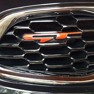 Könnte beinhalten: Nahaufnahme eines schwarzen Kühlergrills mit Chromumrandung. Der Kühlergrill hat ein Wabenmuster und trägt ein orangefarbenes "GT"-Emblem.