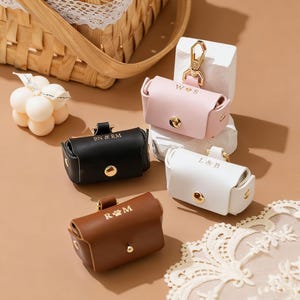 May include: A selection of personalised leather pet waste bag holders in black, brown, pink, and white. Each holder has gold hardware and initials or a paw print. The holders are displayed on a neutral surface.