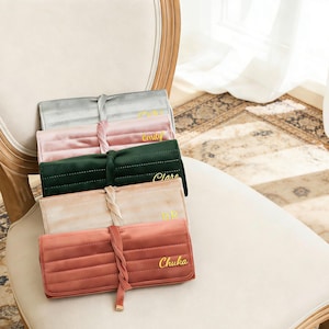 May include: A collection of five velvet cosmetic bags in various colors: gray, pink, green, beige, and coral. Each bag is personalized with a name in gold lettering. The bags are arranged on a white chair.