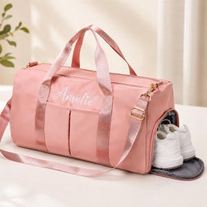 Puede incluir: Una bolsa de deporte rosa polvoriento con el nombre Amelie bordado en letra cursiva blanca en la parte frontal. La bolsa incluye dos asas superiores, una correa de hombro extraíble con herrajes dorados y un compartimento lateral con zapatillas.