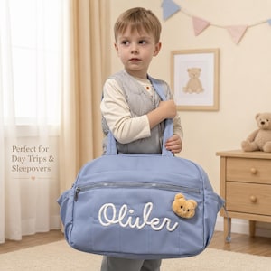 Puede incluir: Una bolsa de viaje azul claro con un bolsillo frontal horizontal con cremallera, una aplicación de felpa con forma de cara de oso color canela y el nombre Oliver bordado en letras cursivas blancas. Incluye dos asas superiores y bolsillos laterales.