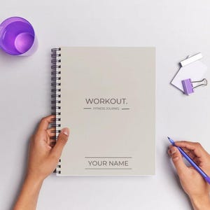 May include: A white spiral bound notebook with the text "WORKOUT. FITNESS JOURNAL" and "YOUR NAME" on the cover. A person is holding the notebook with their left hand and a purple pen with their right hand.