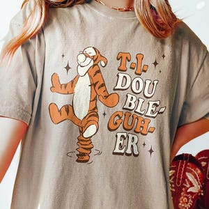 Könnte beinhalten: Khaki T-Shirt mit einer Cartoon-Illustration von Winnie Puuh in einer Tanzpose mit dem Text "T.I. Double-guh-er".