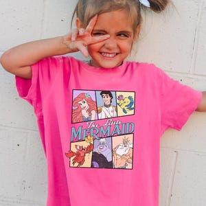 Puede incluir: Una camiseta rosa brillante con un gráfico de La Sirenita de Disney. El diseño incluye a Ariel, el Príncipe Eric, Flounder, Sebastián, Úrsula y el Rey Tritón. La camiseta la lleva una niña pequeña con el pelo recogido en coletas.