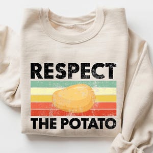 Puede incluir: Sudadera color crema con el texto negro "RESPECT THE POTATO", sobre un gráfico de una patata. El texto está sobre un diseño de rayas horizontales en verde, amarillo y rojo. Las mangas están dobladas.