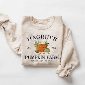 May include: Cream-colored sweatshirt with the text "Hagrid's Pumpkin Farm" and a graphic of pumpkins and leaves. The sweatshirt has rolled-up sleeves and a crew neck. The text includes the year "1962" and the phrase "Magically Grown".