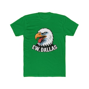 May include: A bright green short-sleeve t-shirt featuring a graphic of a bald eagle head with a yellow beak and a red tongue. Below the eagle, the text EW, DALLAS is printed in a bold white font with a black outline on the chest area.