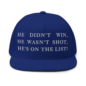 Puede incluir: Gorra de b&eacute;isbol azul real con visera plana. La gorra tiene texto bordado en blanco: "HE DIDN'T WIN, HE WASN'T SHOT, HE'S ON THE LIST!" La parte inferior de la visera es verde.