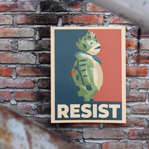 May include: Poster showing a green creature with a blue collar and a red button, set against a blue and red background. The word "RESIST" is printed in bold, white letters at the bottom of the poster.