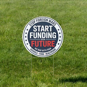 May include: A round white and navy blue sign with the text "STOP FOREIGN WARS START FUNDING OUR FUTURE" in bold letters. The sign also includes the phrases "NO WAR WITH IRAN", "SCHOOLS", "FOOD", and "HEALTHCARE". The sign is on a metal stake.