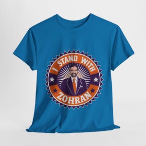 May include: A blue t-shirt with a circular graphic that reads "I STAND WITH ZOHRAN." The graphic includes a man in a suit and tie, stars, and a decorative border. The shirt is made of a soft material and is suitable for casual wear.