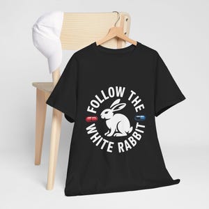 Puede incluir: Camiseta negra con un gráfico blanco de un conejo dentro de un círculo con el texto "FOLLOW THE WHITE RABBIT". El diseño incluye ilustraciones de pastillas rojas y azules. Una gorra blanca descansa sobre una silla de madera.