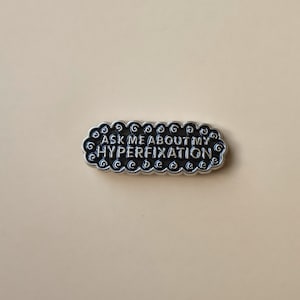 Op de afbeelding: Een zilverkleurige pin met de tekst "ASK ME ABOUT MY HYPERFIXATION" in een zwarte, gestileerde letter. De pin heeft een wolkvorm met decoratieve krullen langs de randen. De achtergrond is effen lichtbruin.