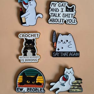 Op de afbeelding: Een verzameling van zes kat-thema emaille pins. De pins tonen verschillende kattenillustraties met humoristische tekst, waaronder "My cat and I talk shit about you" en "Say that again". Het kleurenpalet omvat zwart, wit en accenten van rood en geel.