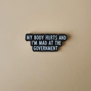 Op de afbeelding: Een zwart-witte rechthoekige sticker met de tekst "MY BODY HURTS AND I'M MAD AT THE GOVERNMENT". De sticker heeft een zwarte rand en staat tegen een beige achtergrond.