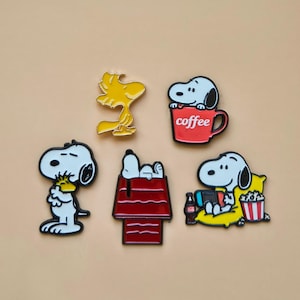Puede incluir: Conjunto de cinco pines esmaltados con personajes de Snoopy y Woodstock. Los diseños incluyen a Snoopy con café, Snoopy en su caseta, Woodstock y Snoopy con palomitas y una bebida. Cada pin tiene un contorno negro y colores vibrantes.