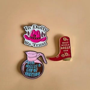 Può includere: Tre spille smaltate a tema musica country. Una spilla presenta un cappello da cowboy rosa con la scritta "In Dolly We Trust". Un'altra spilla è uno stivale da cowboy rosso con la scritta "What Would Dolly Do?" La terza spilla è una caffettiera rosa con la scritta "Pour Myself A Cup Of Ambition."