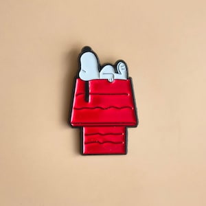 Könnte beinhalten: Eine Metall-Emaille-Anstecknadel, die einen weißen Cartoon-Hund mit schwarzen Ohren zeigt, der auf dem Dach einer leuchtend roten Hundehütte liegt. Das Design ist mit schwarzem Metall umrandet und zeigt den Hund mit Kopf und Pfoten auf dem Dach.