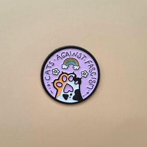 May include: A round, purple Cats Against Fascism sticker with a black border. The design features two cat paws, one orange and white, the other black and white, reaching towards a rainbow. The text "CATS AGAINST FASCISM" is in a circle around the design.