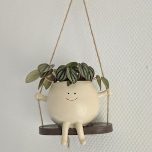 Op de afbeelding: Een grillige, crèmekleurige keramische plantenbak in de vorm van een lachend karakter, compleet met armen, benen en een kleine bruine schommel. De plantenbak bevat een kleine groene plant met patronen op de bladeren. Hij hangt aan een jute touw tegen een witte, getextureerde muur.
