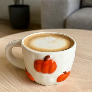 Può includere: Una tazza di ceramica bianca piena di un latte macchiato, con un design decorativo di zucche arancioni. La tazza è su un tavolo di legno chiaro, con uno sfondo sfocato di una pianta e posti a sedere.