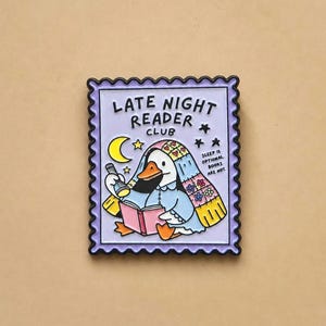 May include: A rectangular enamel pin shaped like a postage stamp with a light purple background. It features a white duck wearing a floral blanket, holding a flashlight and an open pink book. Text reads LATE NIGHT READER CLUB and SLEEP IS OPTIONAL. BOOKS ARE NOT.