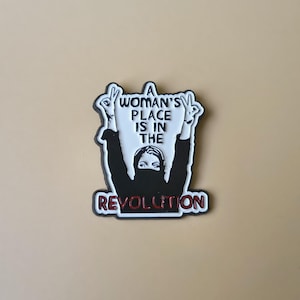 Può includere: Una spilla in smalto bianco e nero raffigurante una persona con le braccia alzate che fa il segno di pace. Il testo recita A WOMAN'S PLACE IS IN THE REVOLUTION, con la parola REVOLUTION in lettere rosse su uno sfondo bianco e bordo nero.