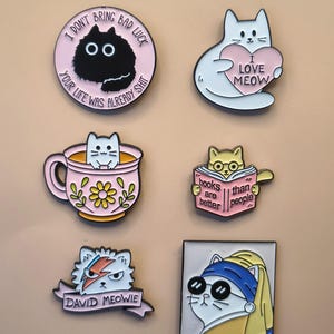 May include: Six enamel pins featuring cat designs. One is a round pink pin with a black cat and text. Another is a white cat holding a pink heart. A third shows a cat in a pink teacup. A fourth is a cat reading a book. A fifth is a cat with a lightning bolt, and the last is a cat wearing a headscarf and sunglasses.