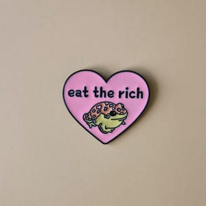 May include: A pink heart-shaped pin with the text "eat the rich" in white. A cartoon frog with yellow and brown spots is in the center. The pin has a black border and is set against a beige background.